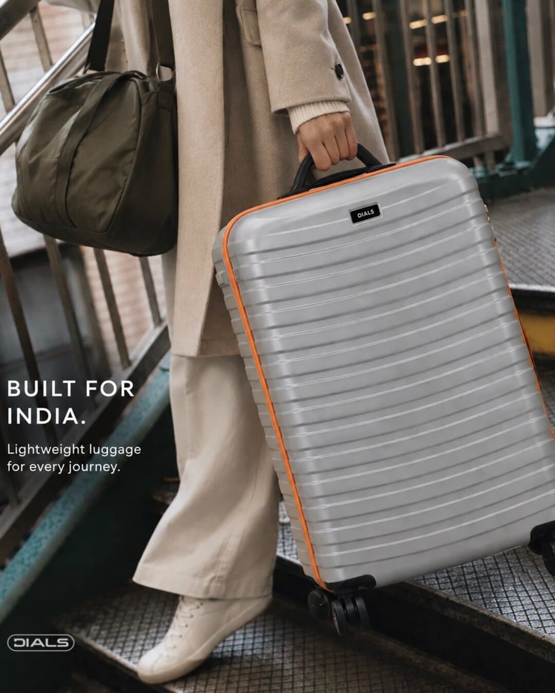 Dials post: “Built for India. Lightweight luggage for every journey.” A traveller carries a silver case with orange trim down a station staircase.